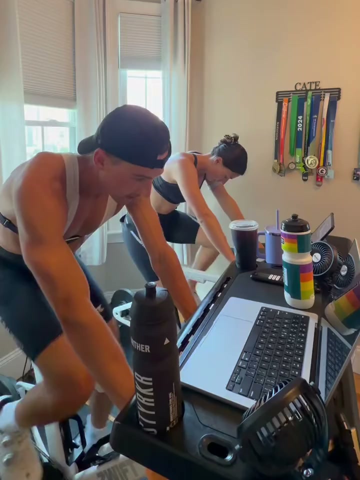 Hank wearing a backward cap while cycling indoors, with both riders and the trainer setup visible.